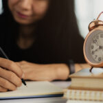 A imagem mostra uma jovem escrevendo em um caderno, com um relógio despertador retrô em destaque sobre uma pilha de livros. O foco está nas mãos dela, sugerindo concentração e organização nos estudos, enquanto o relógio remete à importância da gestão do tempo — aspecto essencial na preparação para provas como o ENEM. A cena transmite disciplina e foco acadêmico.