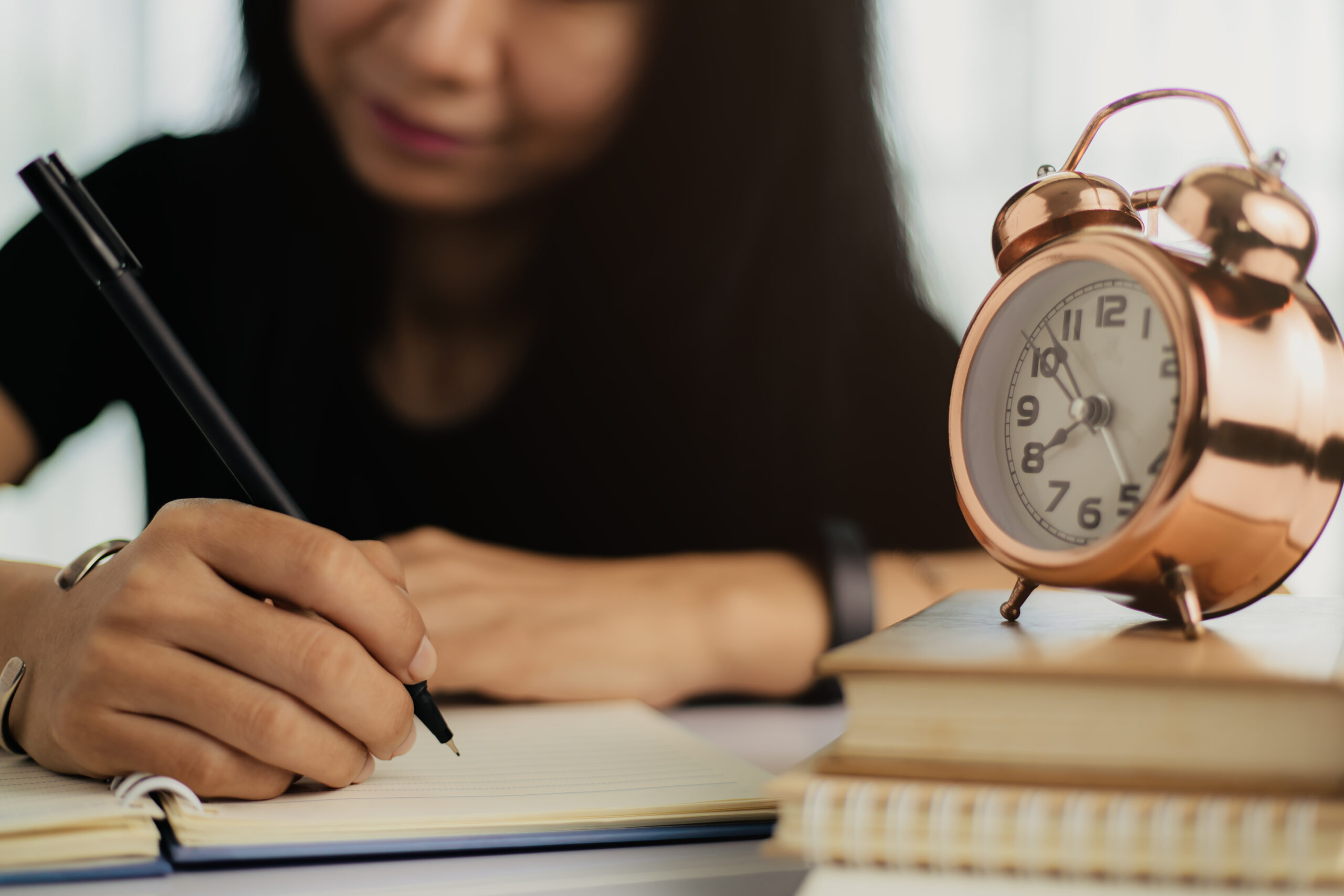 A imagem mostra uma jovem escrevendo em um caderno, com um relógio despertador retrô em destaque sobre uma pilha de livros. O foco está nas mãos dela, sugerindo concentração e organização nos estudos, enquanto o relógio remete à importância da gestão do tempo — aspecto essencial na preparação para provas como o ENEM. A cena transmite disciplina e foco acadêmico.