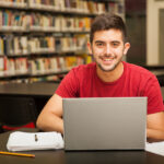 A imagem mostra um jovem sorridente, vestindo uma camiseta vermelha, sentado em uma biblioteca em frente a um notebook. À sua frente, há cadernos, folhas e canetas, sugerindo que ele está estudando ou se preparando para processos seletivos como o Sisu. O ambiente é silencioso e rodeado por estantes cheias de livros, transmitindo uma atmosfera de foco e dedicação aos estudos.
