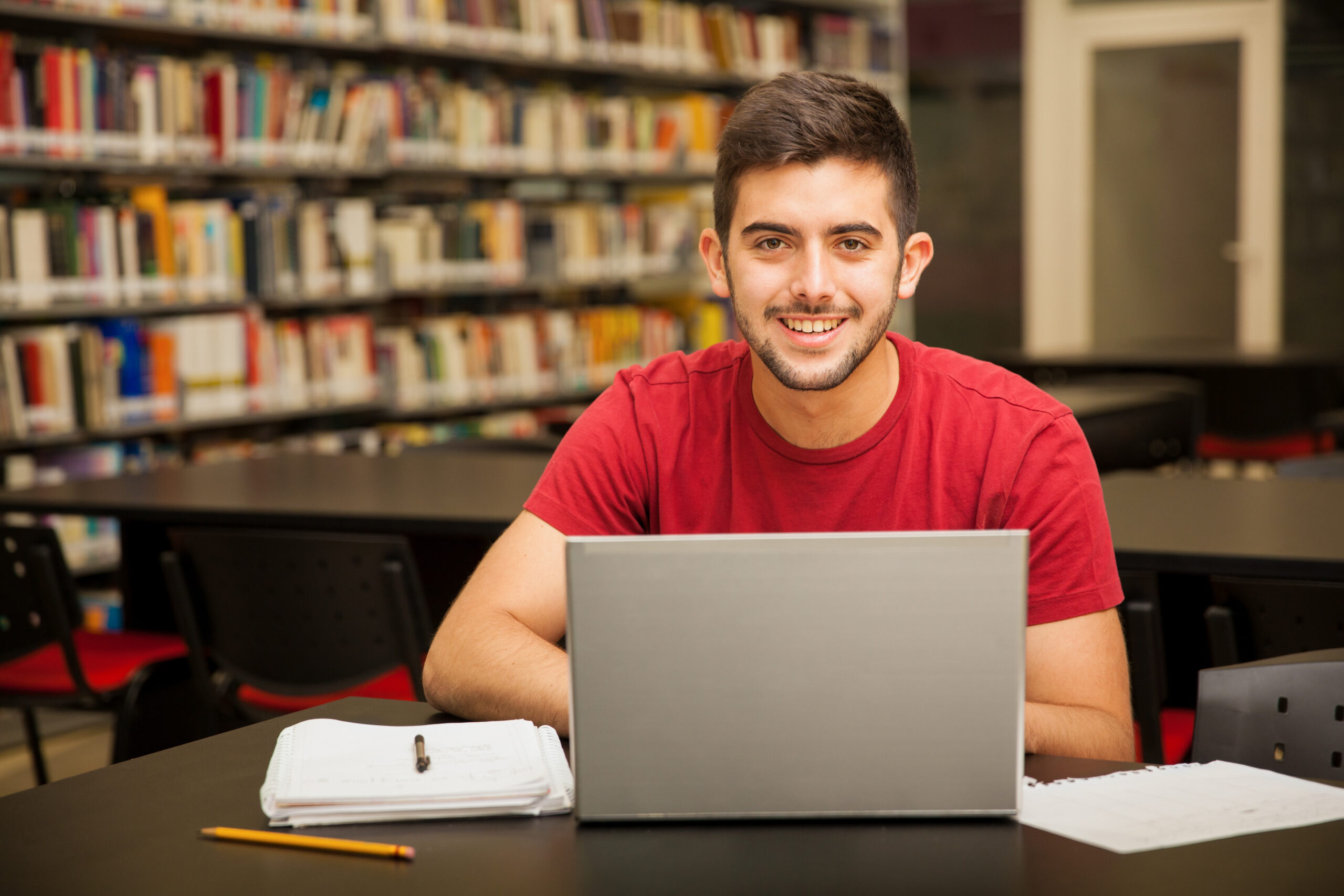 A imagem mostra um jovem sorridente, vestindo uma camiseta vermelha, sentado em uma biblioteca em frente a um notebook. À sua frente, há cadernos, folhas e canetas, sugerindo que ele está estudando ou se preparando para processos seletivos como o Sisu. O ambiente é silencioso e rodeado por estantes cheias de livros, transmitindo uma atmosfera de foco e dedicação aos estudos.