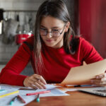 Jovem com óculos e blusa vermelha estudando em uma mesa com papéis, livros, laptop e canetas, em um ambiente doméstico com cozinha ao fundo.