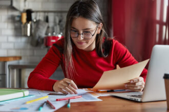 Jovem com óculos e blusa vermelha estudando em uma mesa com papéis, livros, laptop e canetas, em um ambiente doméstico com cozinha ao fundo.