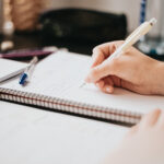 O ChatGPT disse: Pessoa escrevendo à mão em um caderno espiral sobre uma mesa, com canetas e papéis ao redor, em ambiente de estudo ou trabalho.