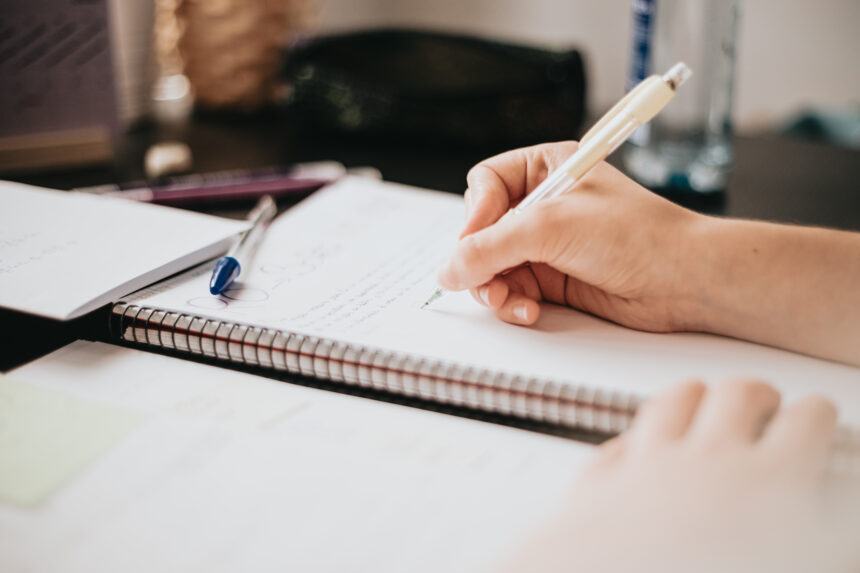 O ChatGPT disse: Pessoa escrevendo à mão em um caderno espiral sobre uma mesa, com canetas e papéis ao redor, em ambiente de estudo ou trabalho.