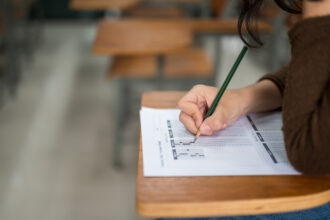 O ChatGPT disse: Pessoa preenchendo um cartão de respostas com lápis durante prova em sala de aula, em foco o movimento da mão e o formulário.