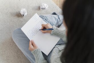 Uma jovem aluna parece concentrada e ansiosa enquanto escreve em um caderno, possivelmente estudando para a redação do ENEM