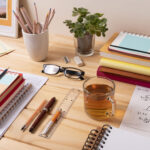 Mesa de estudos organizada com cadernos, livros empilhados, papéis com fórmulas matemáticas, lápis de cor em um copo, óculos, calculadora, canetas, régua, um copo de chá e uma planta suculenta em vaso de vidro.